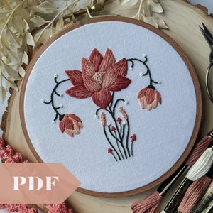 May include: Embroidered hoop art featuring a pink and red flower with green stems and leaves. The hoop is made of wood and has a white fabric background.