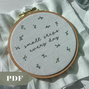 Op de afbeelding: Een witte borduurring met een houten frame. De stof in de ring heeft de zin "small steps every day" geborduurd in zwarte draad. Er zijn kleine groene geborduurde bladeren verspreid rond de zin.