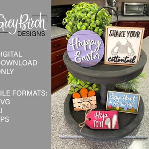 May include: A two-tiered wooden tray with a variety of Easter-themed decorations. The top tier features a purple circle with the text "Happy Easter" and a white sign with the text "Shake Your Cottontail". The bottom tier features a sign with the text "Egg Hunt This Way", a sign with the text "Carrots 25¢", and a tag with the text "Hop To It".