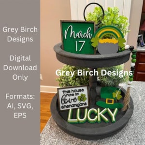 Puede incluir: Una bandeja de madera de dos niveles con un arcoíris verde y dorado, un pote de oro, un trébol, un sombrero de duende y letreros que dicen "March 17", "This house runs on love & shenanigans", y "Lucky".