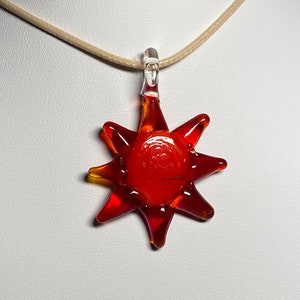 Può includere: Una collana con un ciondolo a forma di sole in vetro rosso. Il sole ha una forma a stella con un centro rosso e bordi arancioni.
