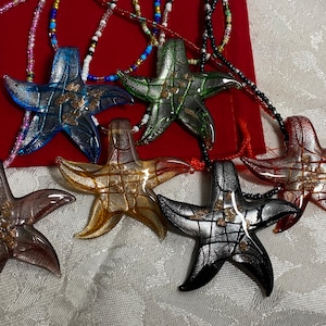 Peut inclure: Six pendentifs en forme d'étoile de mer en verre avec des paillettes dorées, chacun sur une chaîne perlée. Les étoiles de mer sont de différentes couleurs, notamment bleu, vert, marron, rouge et noir.