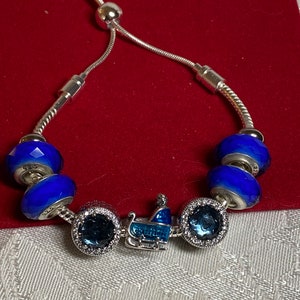 Pode incluir: Um bracelete de prata com contas de vidro azul e transparente e um amuleto de prata de um trenó com um design azul e branco.