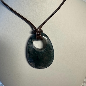 May include: A dark green moss agate pendant with a smooth, polished, oval shape featuring a circular cutout at the top. The pendant is suspended from a simple, dark brown braided cord necklace that loops through the opening of the stone.
