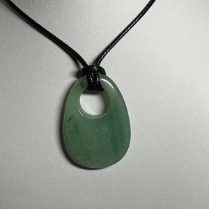 May include: A smooth, translucent sage green stone pendant with a circular cutout at the top, suspended from a simple black cord. The pendant has an organic, rounded teardrop shape with subtle variations in the green color throughout the stone.