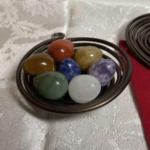 May include: Seven smooth, polished gemstone eggs in a wire nest. The eggs are red, brown, blue, green, white, purple, and yellow.