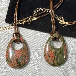 May include: Two teardrop-shaped pendants made of polished unakite stone featuring mottled green and pink patterns. Each pendant is suspended from a brown braided cord necklace with a gold-toned metal lobster clasp and an adjustable chain link.