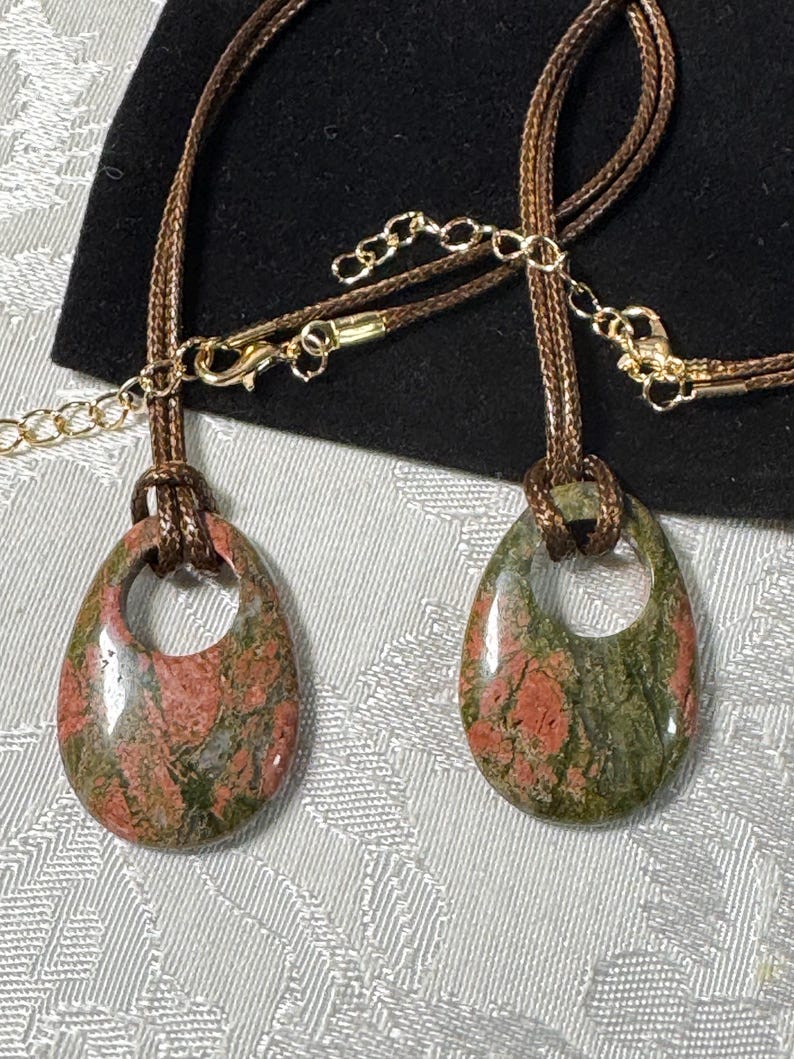 May include: Two teardrop-shaped pendants made of polished unakite stone featuring mottled green and pink patterns. Each pendant is suspended from a brown braided cord necklace with a gold-toned metal lobster clasp and an adjustable chain link.