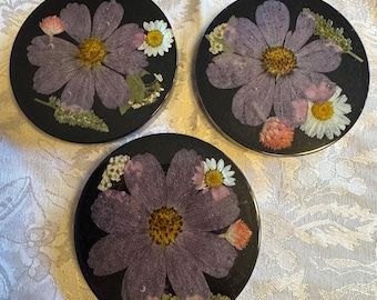 May include: Three round black coasters, each featuring a pressed purple flower with a yellow center, surrounded by smaller white and pink flowers and green foliage. The coasters are set on a white patterned surface.