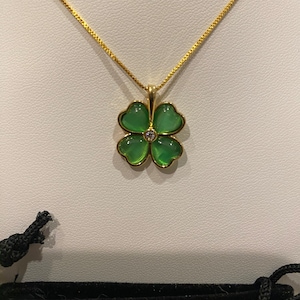 Puede incluir: Un collar dorado con un colgante de trébol de cuatro hojas. El trébol tiene cuatro hojas verdes en forma de corazón y una pequeña gema transparente en el centro. El collar se muestra sobre una superficie blanca.