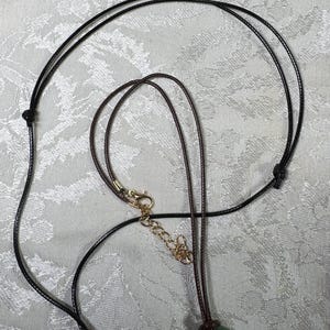 May include: A dark brown cord necklace featuring a circular green stone pendant. The necklace includes a gold-toned metal lobster claw clasp and an adjustable chain extender, allowing for a customizable fit around the neck for various wearers.