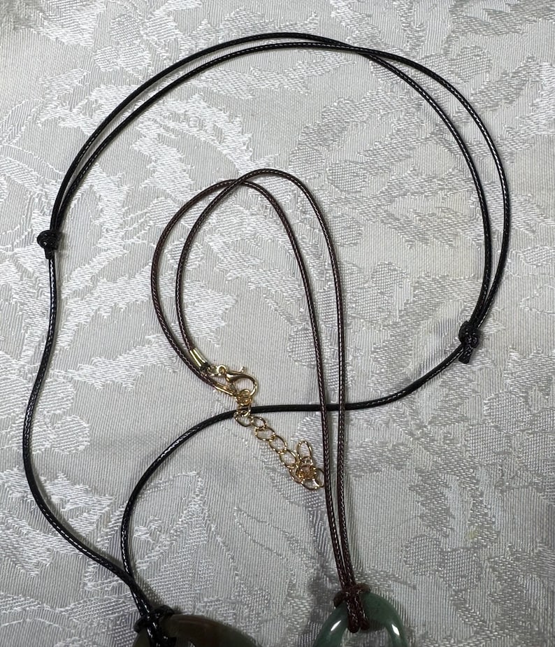 May include: A dark brown cord necklace featuring a circular green stone pendant. The necklace includes a gold-toned metal lobster claw clasp and an adjustable chain extender, allowing for a customizable fit around the neck for various wearers.