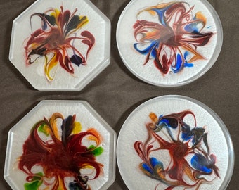 Puede incluir: Cuatro posavasos con fondo blanco y diseños abstractos coloridos. Los diseños son remolinos de pintura roja, azul, amarilla y verde.