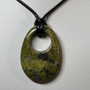 May include: A polished, oval-shaped green stone pendant with a circular cutout at the top. The stone features mottled patterns of olive, lime, and dark charcoal gray. It is suspended from a simple, thin black cord that loops through the opening.