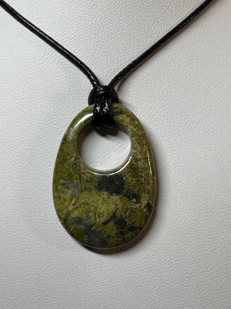 May include: A polished, oval-shaped green stone pendant with a circular cutout at the top. The stone features mottled patterns of olive, lime, and dark charcoal gray. It is suspended from a simple, thin black cord that loops through the opening.