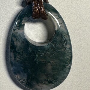 May include: A teardrop-shaped moss agate pendant featuring deep green, fern-like inclusions within a translucent, polished stone. The pendant has a circular cutout at the top and is suspended from a dark brown, braided cord necklace.