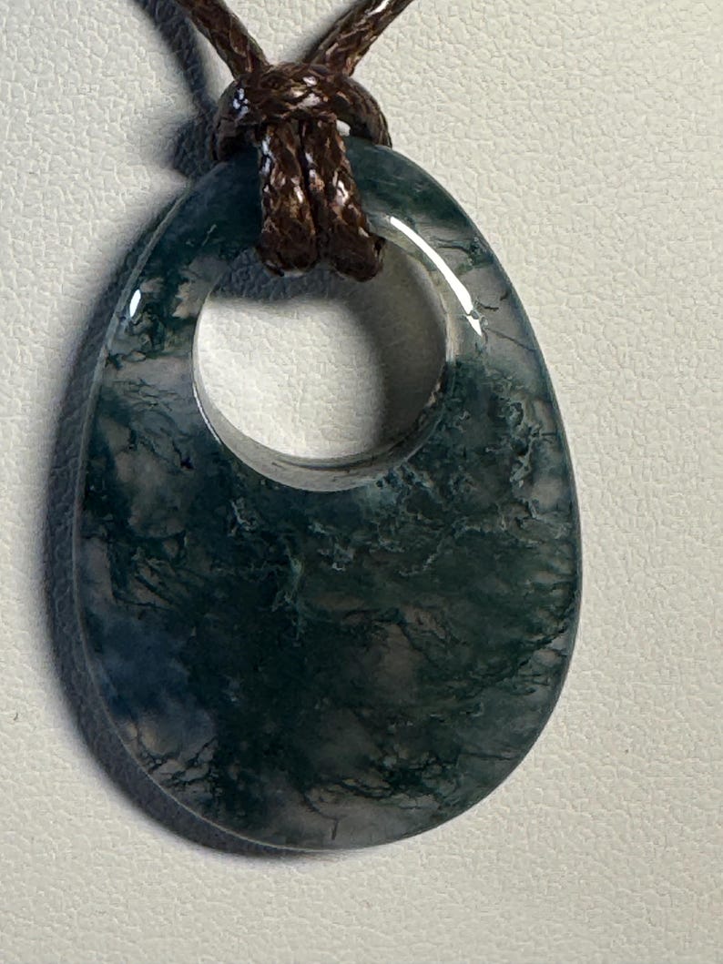 May include: A teardrop-shaped moss agate pendant featuring deep green, fern-like inclusions within a translucent, polished stone. The pendant has a circular cutout at the top and is suspended from a dark brown, braided cord necklace.