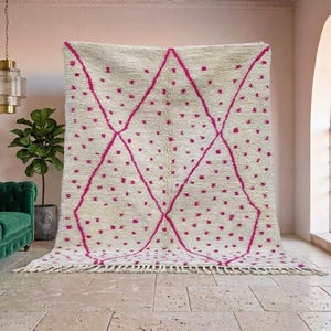 May include: A large, cream-colored rug with a geometric design. The rug features a pattern of pink diamonds and scattered pink dots. The rug has a fringe along the bottom edge. This rug would be a great addition to any living room.