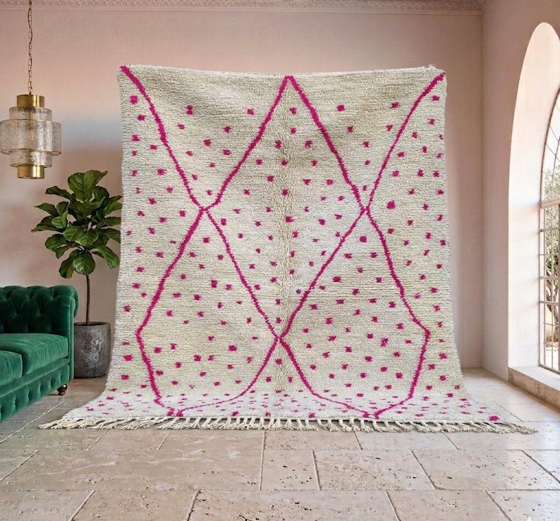 May include: A large, cream-colored rug with a geometric design. The rug features a pattern of pink diamonds and scattered pink dots. The rug has a fringe along the bottom edge. This rug would be a great addition to any living room.