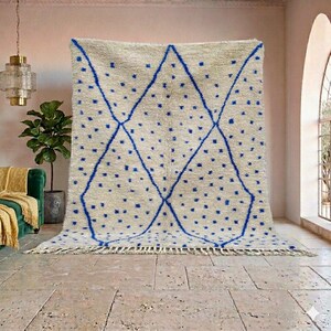 May include: A large, cream-colored rug with a geometric design. The rug features a pattern of blue diamonds and scattered blue dots. The rug has a fringe along the bottom edge and is displayed against a neutral wall.