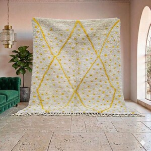 May include: A large, off-white rug with a geometric design. The rug features a repeating pattern of yellow diamonds and small yellow dots. The rug has a fringe along the bottom edge. The rug is displayed indoors.