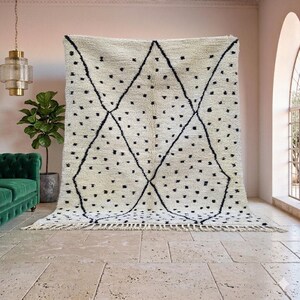 May include: A large, cream-colored rug with a geometric design. The rug features a diamond pattern outlined in black, with scattered black dots. The rug has a fringed edge and is displayed against a pink wall.