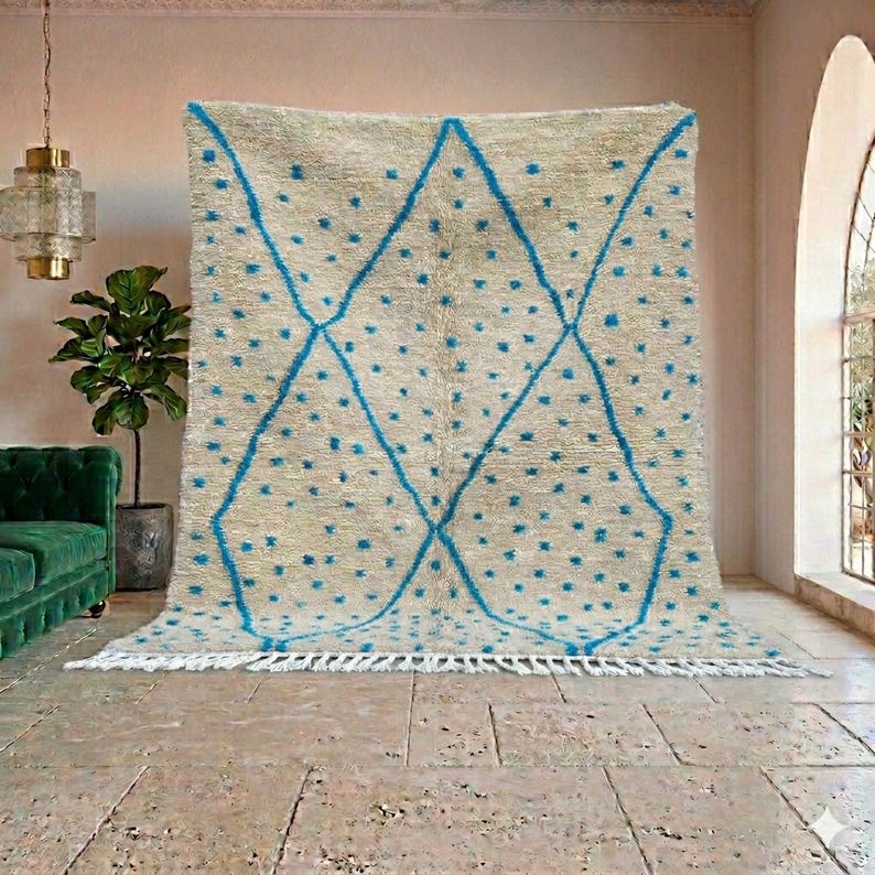 May include: A large, beige rug with a geometric design. The rug features a pattern of blue diamonds and scattered blue dots. The rug has a fringe along the bottom edge and is displayed in a room with a green sofa and a potted plant.