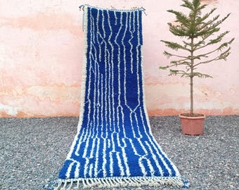 May include: A blue and white patterned rug with a fringe. The rug features a geometric design with vertical white lines on a blue background. The rug is displayed against a pink wall and a small potted tree.
