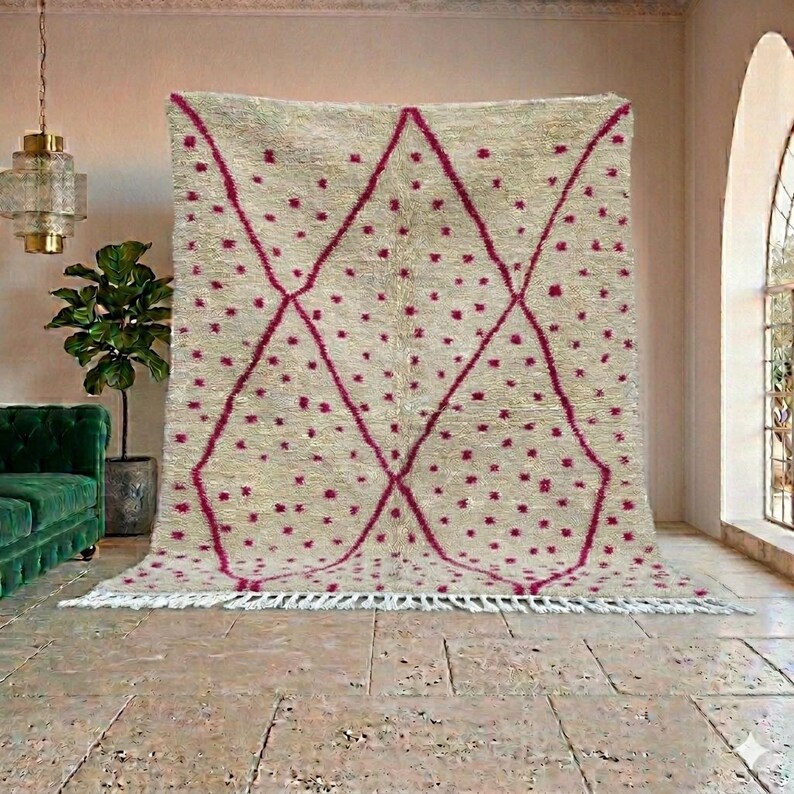 May include: A large, beige rug with a geometric design. The rug features a pattern of magenta diamonds and scattered magenta dots. The rug has a fringe along the bottom edge and is displayed in a room with a green sofa and a potted plant.