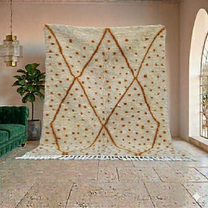 May include: A large, cream-colored rug with a geometric diamond pattern in a warm brown hue. The rug features small, scattered brown dots and a fringe along the bottom edge. The rug is displayed in a room with a green sofa and a potted plant.