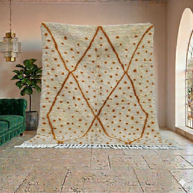May include: A large, cream-colored rug with a geometric diamond pattern in a warm brown hue. The rug features small, scattered brown dots and a fringe along the bottom edge. The rug is displayed in a room with a green sofa and a potted plant.