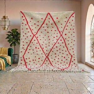 May include: A large, beige rug with a geometric red design. The rug features a diamond pattern with red dots and lines. The rug has a fringed edge and is displayed in a room with a neutral color palette.