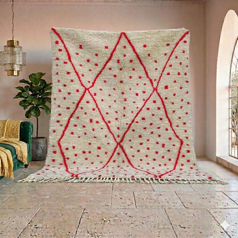 May include: A large, beige rug with a geometric red design. The rug features a diamond pattern with red dots and lines. The rug has a fringed edge and is displayed in a room with a neutral color palette.