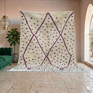 May include: A large, beige rug with a geometric pattern of purple diamonds and scattered dots. The rug has a fringe along the bottom edge and is displayed in a room with a green sofa and a potted plant.
