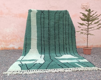 May include: A green and white patterned rug with black vertical lines and a fringed edge. The rug is displayed outdoors with a small potted tree in the background. The rug's design features geometric shapes.