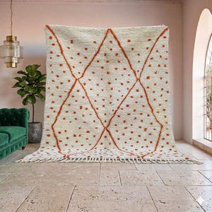 May include: Cream-colored rug with a geometric design. The rug features a diamond pattern in a burnt orange color, with small orange dots scattered across the surface. The rug has a fringed edge.