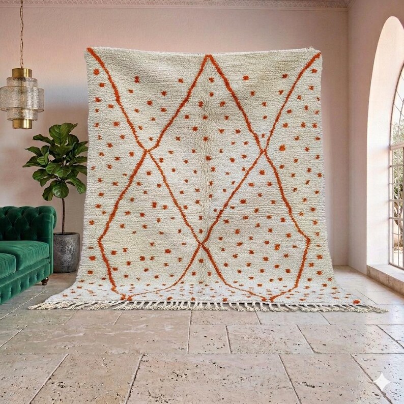 May include: Cream-colored rug with a geometric design. The rug features a diamond pattern in a burnt orange color, with small orange dots scattered across the surface. The rug has a fringed edge.