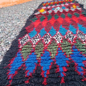Puede incluir: Una colorida alfombra rectangular con un patrón geométrico. La alfombra presenta un diseño de diamantes en rojo, azul, verde y blanco, con una franja negra. La alfombra está hecha de material tejido y es adecuada para la decoración del hogar.