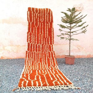 Op de afbeelding: Een rechthoekig tapijt met een oranje en wit geometrisch patroon. Het tapijt heeft een getextureerd oppervlak en een gefranjerde rand. Het wordt buiten gepresenteerd op een bed van kleine grijze stenen.