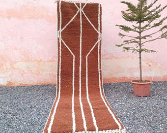 May include: A rectangular brown rug with a geometric white design. The rug has a series of vertical white lines and a diamond-shaped pattern at the top. The rug has a fringe at the bottom. The rug is approximately 152 cm long.