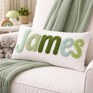May include: A rectangular white throw pillow featuring the name James in large, textured, three-dimensional letters. The letters are crafted in varying shades of green, including light mint, sage, and dark forest green, with a tufted finish.