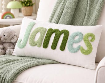Op de afbeelding: Een rechthoekig wit sierkussen met de naam James in verhoogde, getufte letters met textuur. De letters wisselen af tussen tinten licht muntgroen, saliegroen en donker bosgroen, wat zorgt voor een zachte en tactiele uitstraling.