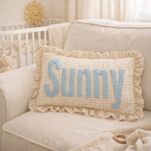May include: A rectangular throw cushion featuring a beige and white gingham pattern with a ruffled border. The word Sunny is embroidered in light blue, textured, raised lettering across the centre of the cushion, which is placed on a sofa.