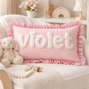 Könnte beinhalten: Ein rechteckiges Kissen mit rosa-weißem Vichy-Muster und Rüschenrand. In der Mitte steht der Name violet in weißen, strukturierten, dreidimensionalen Buchstaben. Ein beiger Plüschhase und eine Holzperlengirlande liegen vor dem Kissen.