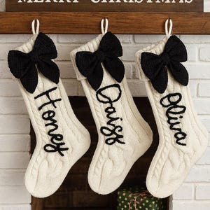 Op de afbeelding: Drie crèmekleurige kerstsokken met zwarte strikken en namen in het zwart geborduurd: Honey, Daisy en Olivia. Ze hangen aan een houten plank met de tekst "MERRY CHRISTMAS".
