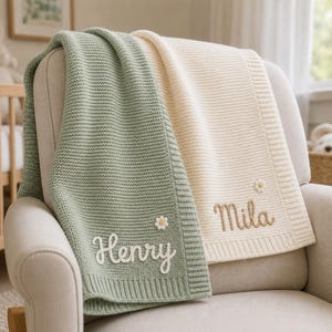 Könnte beinhalten: Eine salbeigrüne und cremefarbene Strickdecke mit geripptem Rand. Der salbeigrüne Teil ist mit dem Namen Henry in weißer Schreibschrift bestickt, der cremefarbene Teil mit dem Namen Mila in brauner Schreibschrift, jeweils mit einer kleinen Blume.