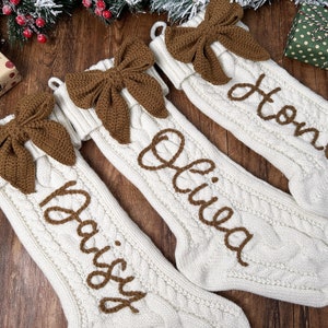 May include: Three white knitted Christmas stockings with brown crocheted bows and personalised names: "Honey", "Olivia", and "Daisy".