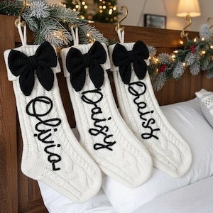 May include: Three cream-colored cable knit Christmas stockings, each featuring a large black knit bow at the top cuff. The stockings are personalized with black embroidered script text reading Olivia, Daisy, and Daisy, hanging from gold hooks.