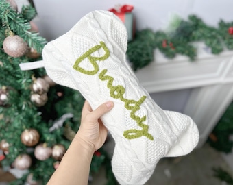 May include: A white cable-knit Christmas stocking shaped like a dog bone. The stocking features the name Buddy embroidered in green yarn across the front. It includes a white fabric loop at the top for hanging and has a textured knit pattern.