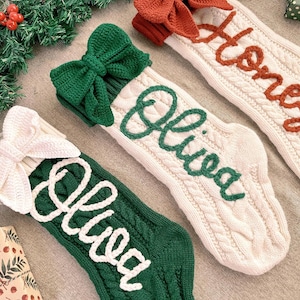 Peut inclure: Trois chaussettes de Noël en tricot avec des textures torsadées et de grands nœuds sur le dessus. Une chaussette verte a un nœud blanc et le nom Olivia, une blanche a un nœud vert et le nom Olivia, et une blanche a un nœud rouille et le nom Honey.